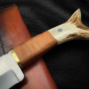 May include: A hunting knife with a brown leather sheath and a stag horn handle. The blade is made of stainless steel and has a polished finish.