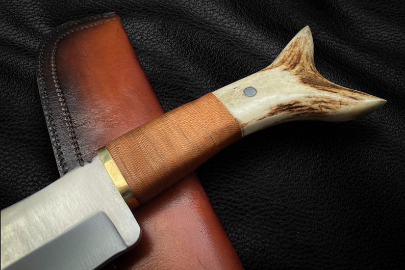 May include: A hunting knife with a brown leather sheath and a stag horn handle. The blade is made of stainless steel and has a polished finish.