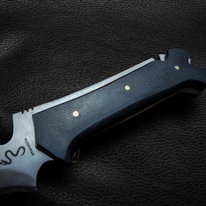 May include: A black and silver knife with a black handle and a silver blade. The handle has a textured surface and three brass accents. The blade has the word "Snakebite" engraved on it.