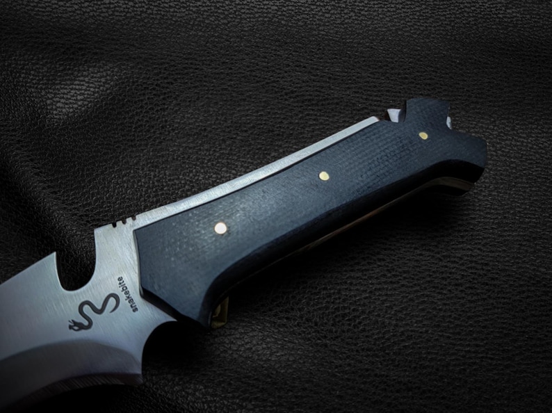 May include: A black and silver knife with a black handle and a silver blade. The handle has a textured surface and three brass accents. The blade has the word "Snakebite" engraved on it.