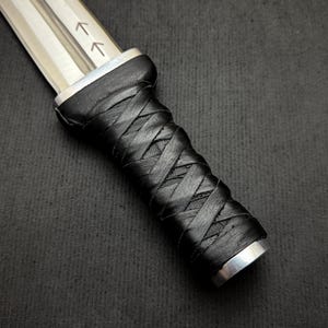 May include: Close-up of a dagger with a silver blade and a black, intricately wrapped leather handle. The blade has two etched arrows. The handle is secured with silver metal at both ends.