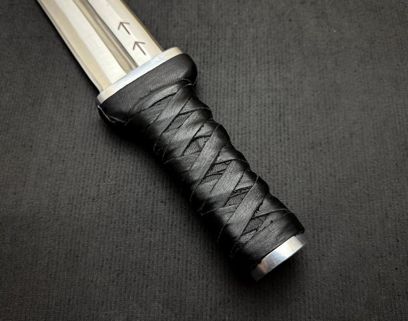 May include: Close-up of a dagger with a silver blade and a black, intricately wrapped leather handle. The blade has two etched arrows. The handle is secured with silver metal at both ends.
