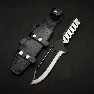 Puede incluir: Un cuchillo táctico negro y plateado con una hoja parcialmente dentada y un mango texturizado. El cuchillo se combina con una funda de cuero negro con un patrón de piel de cocodrilo, asegurada con remaches plateados.