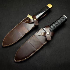 May include: Two handcrafted knives with sheaths. The blades are sheathed in brown leather with gold stitching. The handles are black and wrapped with a textured grip. The sheaths have a leather strap with a metal button closure.