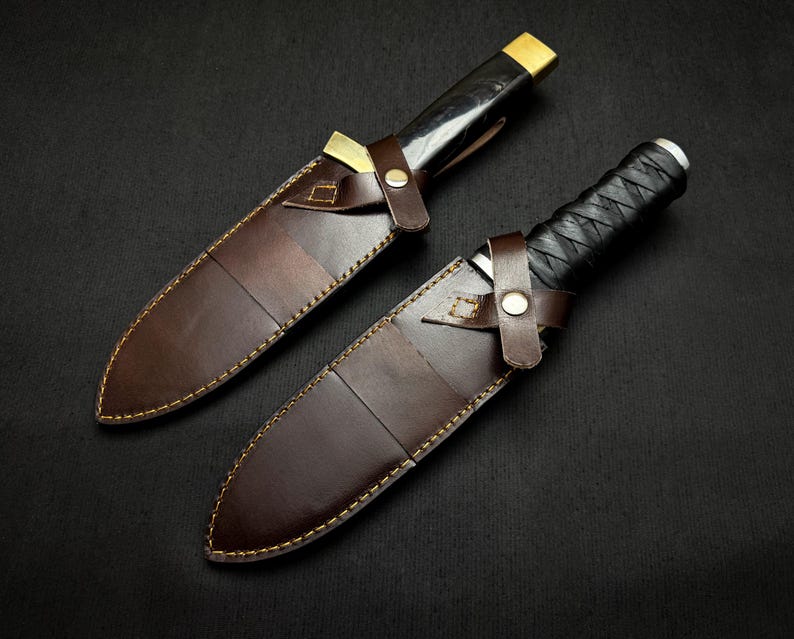 May include: Two handcrafted knives with sheaths. The blades are sheathed in brown leather with gold stitching. The handles are black and wrapped with a textured grip. The sheaths have a leather strap with a metal button closure.