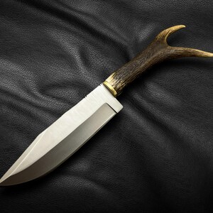 May include: A hunting knife with a brown antler handle and a silver blade. The knife is lying on a black leather surface.