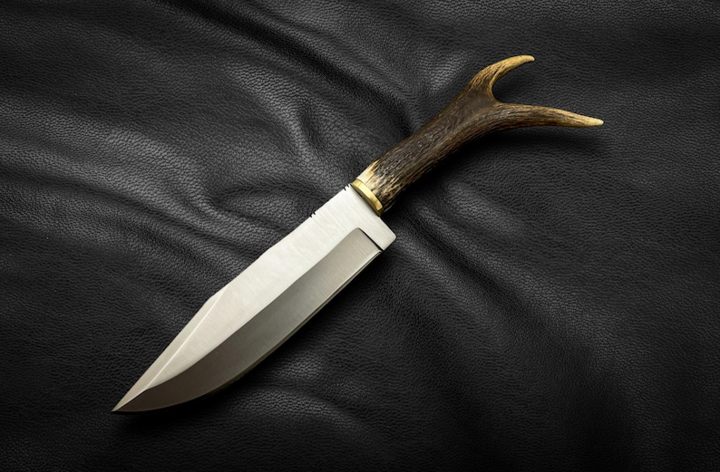 May include: A hunting knife with a brown antler handle and a silver blade. The knife is lying on a black leather surface.