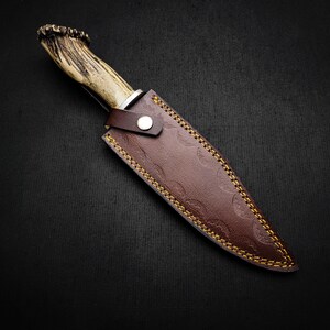May include: A brown leather knife sheath with a decorative stitching pattern and an antler handle knife.