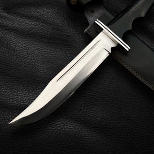 May include: A silver hunting knife with a black leather sheath. The knife has a long, sharp blade and a wooden handle.