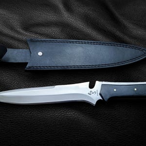 May include: A black leather sheath with a silver knife with a black handle. The knife has a serrated edge and a small hole near the handle.