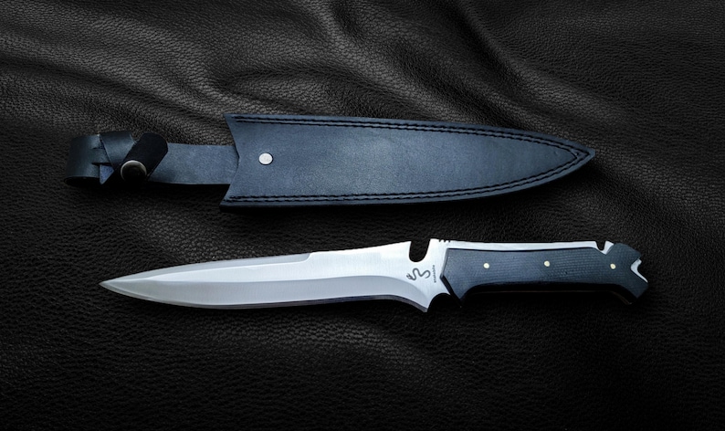 May include: A black leather sheath with a silver knife with a black handle. The knife has a serrated edge and a small hole near the handle.