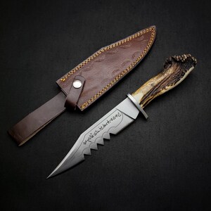 May include: A hunting knife with a brown leather sheath and a stag antler handle. The blade is engraved with Arabic script.