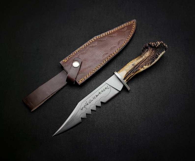 May include: A hunting knife with a brown leather sheath and a stag antler handle. The blade is engraved with Arabic script.