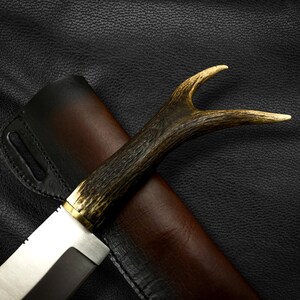 May include: A hunting knife with a brown antler handle and a leather sheath. The knife has a silver blade and a brass accent.