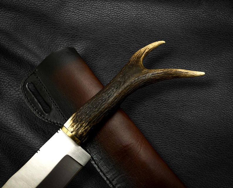 May include: A hunting knife with a brown antler handle and a leather sheath. The knife has a silver blade and a brass accent.