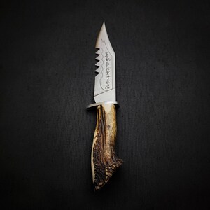 May include: A silver hunting knife with a serrated blade and a stag antler handle. The blade has an inscription that reads "Never Surrender".