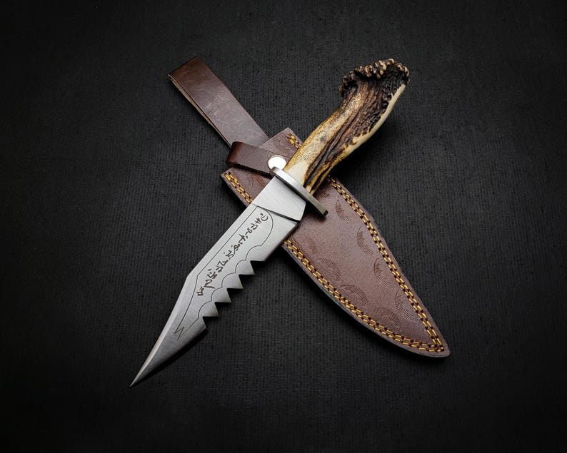 May include: A hunting knife with a brown leather sheath and a stag antler handle. The blade has a serrated edge and is engraved with Arabic script.