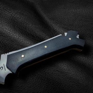 May include: A black and silver knife with a black handle and gold accents. The knife has a serrated blade and a bottle opener. The knife is on a black leather surface. The knife has the word "Snakebite" engraved on the blade.