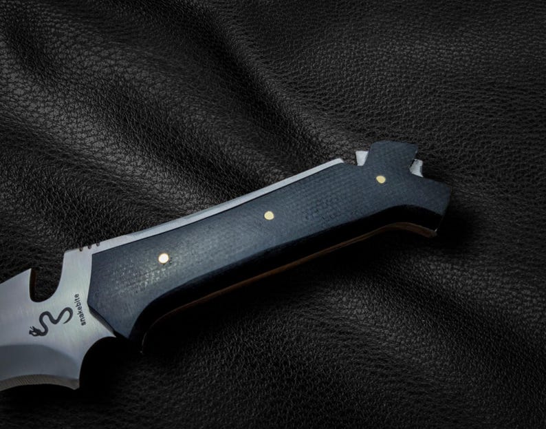 May include: A black and silver knife with a black handle and gold accents. The knife has a serrated blade and a bottle opener. The knife is on a black leather surface. The knife has the word "Snakebite" engraved on the blade.