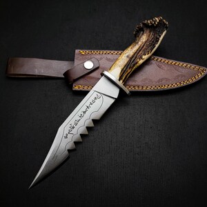 May include: A hunting knife with a brown leather sheath and a stag antler handle. The blade is serrated and has Arabic script engraved on it.