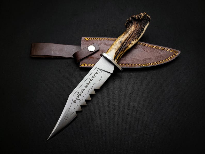 May include: A hunting knife with a brown leather sheath and a stag antler handle. The blade is serrated and has Arabic script engraved on it.