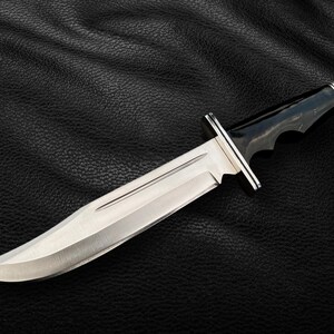 May include: A silver hunting knife with a black handle and a serrated edge. The knife is lying on a black leather surface.