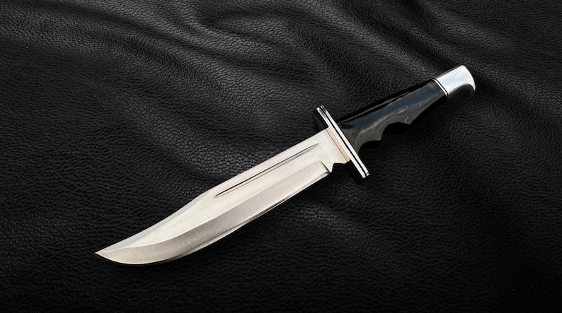 May include: A silver hunting knife with a black handle and a serrated edge. The knife is lying on a black leather surface.