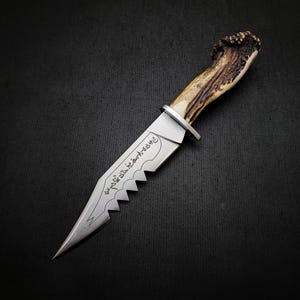 May include: A silver knife with a serrated edge and a brown antler handle. The blade is engraved with the text "the 7th sister art knife".