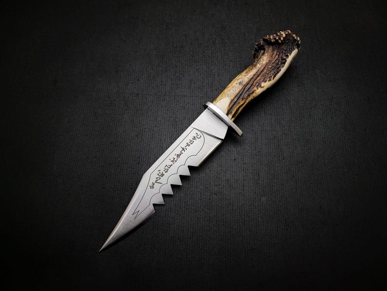 May include: A silver knife with a serrated edge and a brown antler handle. The blade is engraved with the text "the 7th sister art knife".