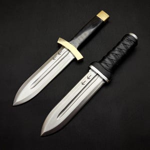 May include: Two daggers with silver blades and black handles. One dagger has a gold crossguard and a black handle. The other dagger has a black wrapped handle. Both daggers have arrows etched on the blade.