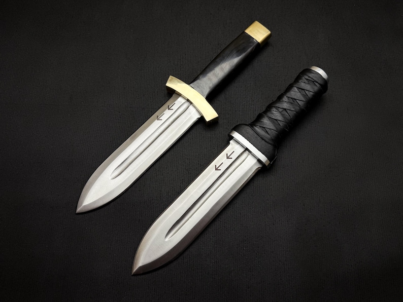 May include: Two daggers with silver blades and black handles. One dagger has a gold crossguard and a black handle. The other dagger has a black wrapped handle. Both daggers have arrows etched on the blade.