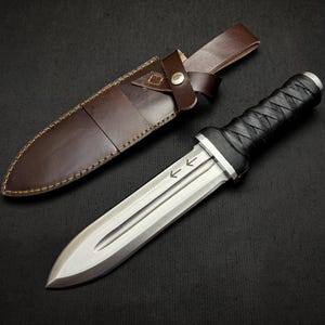 May include: A dagger with a silver blade and a black, wrapped handle, next to a brown leather sheath. The blade has etched arrows. The sheath is brown leather with a strap and button closure. The dagger is a handmade item.