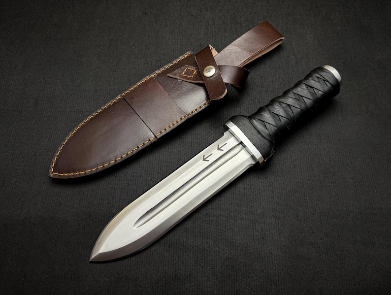May include: A dagger with a silver blade and a black, wrapped handle, next to a brown leather sheath. The blade has etched arrows. The sheath is brown leather with a strap and button closure. The dagger is a handmade item.