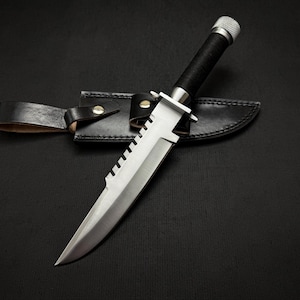 May include: A silver knife with a black handle and a serrated blade. The knife is in a black leather sheath.