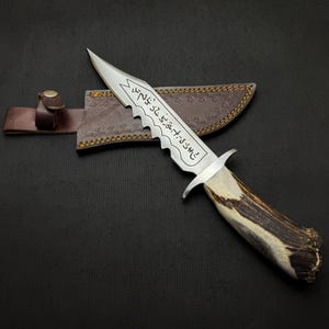 May include: A hunting knife with a serrated blade and a handle made of antler. The blade has etched text. The knife rests on a brown leather sheath with a button closure. The background is black.