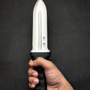 May include: A silver-colored dagger with a pointed tip and a black, textured handle. The handle is wrapped in a crisscross pattern. Two arrows are etched on the blade. The dagger is held in a hand.