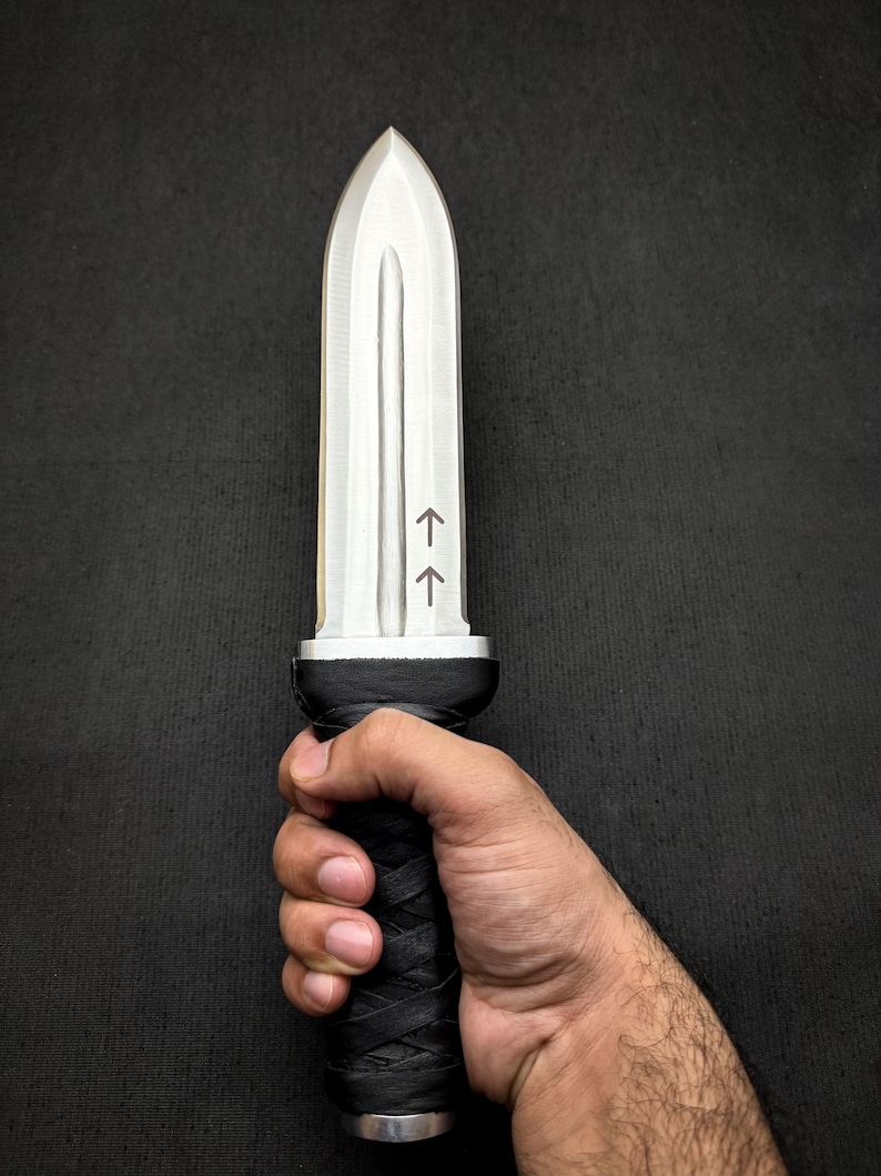 May include: A silver-colored dagger with a pointed tip and a black, textured handle. The handle is wrapped in a crisscross pattern. Two arrows are etched on the blade. The dagger is held in a hand.