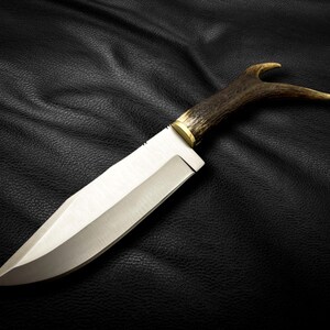 May include: A hunting knife with a long, silver blade and a brown antler handle. The knife is lying on a black leather surface.