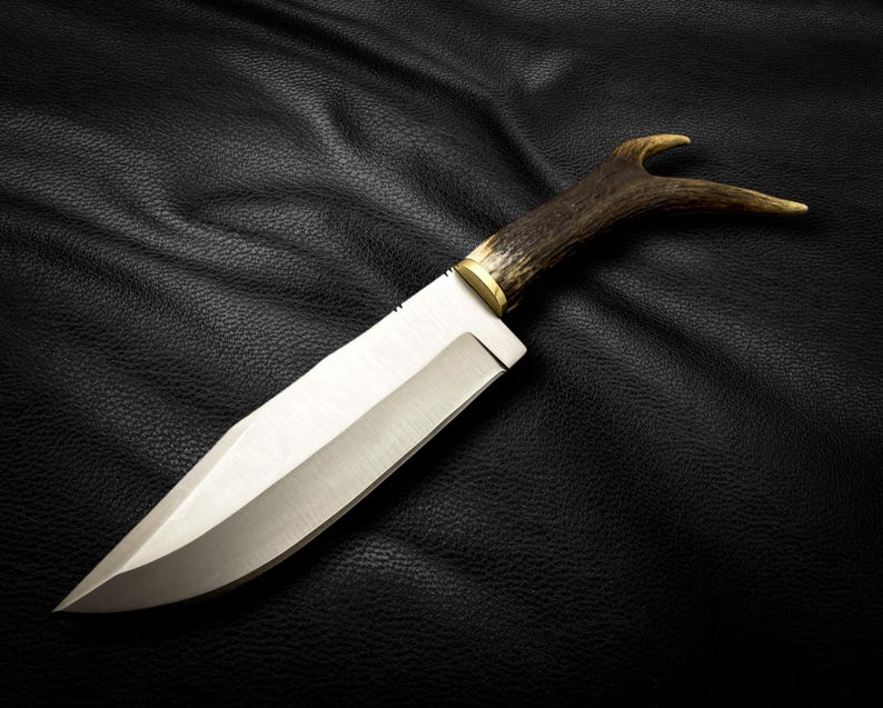 May include: A hunting knife with a long, silver blade and a brown antler handle. The knife is lying on a black leather surface.