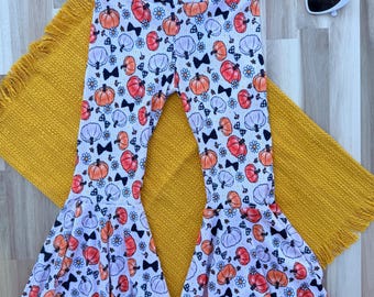 May include: White ribbed bell-bottom pants featuring a repeating pattern of orange pumpkins, white pumpkins, small blue flowers, and black bows. The pants have a high-waisted fit and flared legs. White heart-shaped sunglasses are placed near the top.