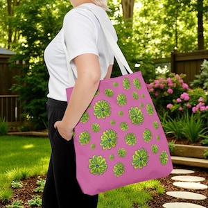 May include: A pink tote bag with a repeating pattern of green and yellow flowers. The bag has white straps and is being carried outdoors. The flowers have a blue center.