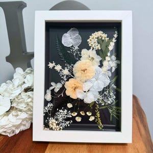 Handmade pressed flower frame with delicate white and beige blossoms arranged on a dark background in a sleek white frame. Minimalist botanical wall art designed in a neutral color palette, perfect for Japandi, Scandinavian, or modern interiors. This dried flower frame can be displayed on walls or shelves, adding a calming natural touch to any space. A timeless home accent and a meaningful gift idea for weddings, anniversaries, housewarmings, or for nature lovers who appreciate simple elegance.