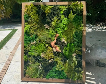 May include: A rectangular framed wall art piece filled with various shades of green moss and ferns. A light brown, branch-like element is placed in the centre of the moss. The frame is made of light brown wood.