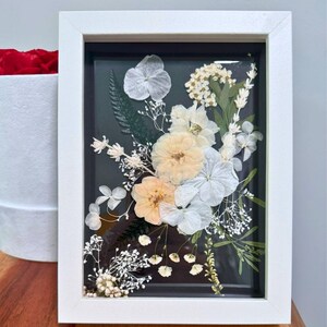 May include: A rectangular white frame containing a pressed floral arrangement on a dark background. The composition features light peach and white flowers, including hydrangea petals, small white blossoms, and green fern leaves behind glass.