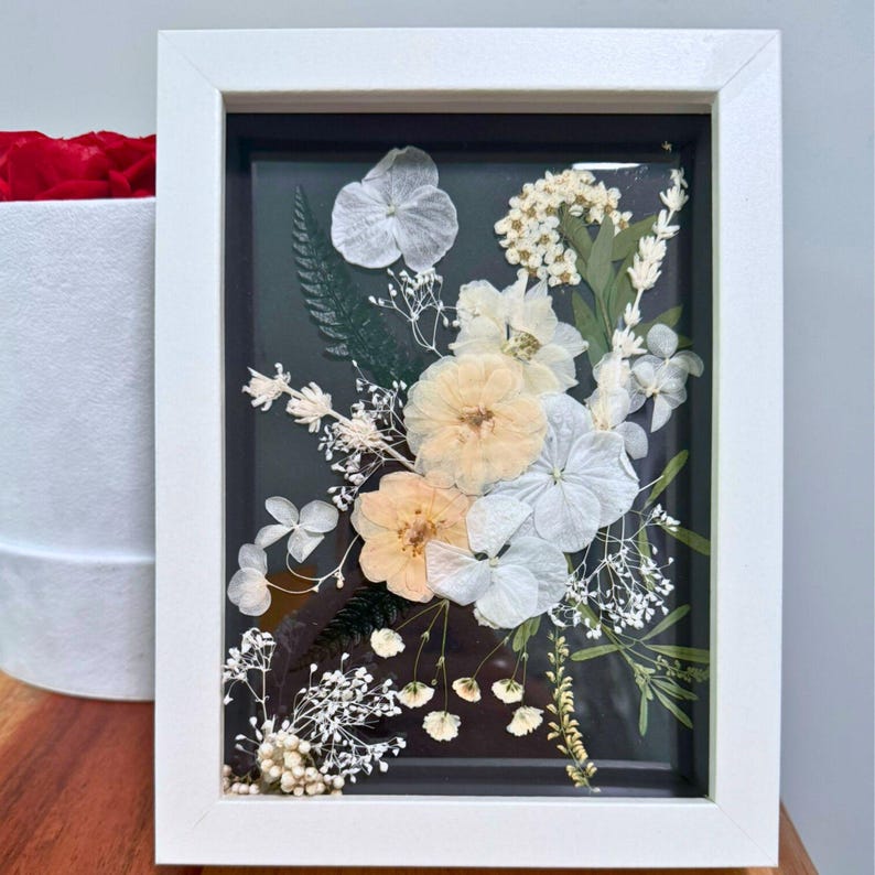 May include: A rectangular white frame containing a pressed floral arrangement on a dark background. The composition features light peach and white flowers, including hydrangea petals, small white blossoms, and green fern leaves behind glass.