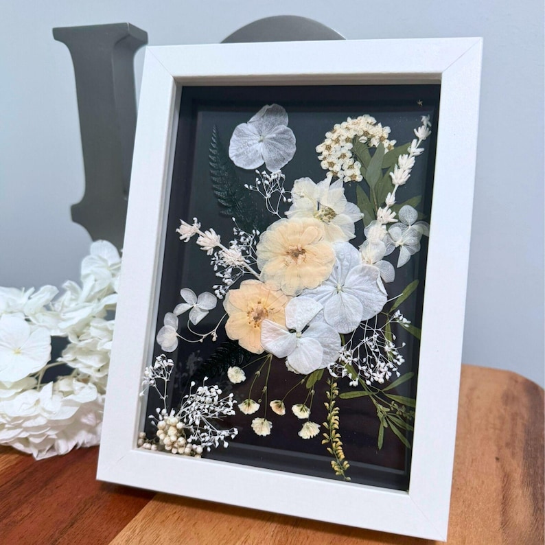 May include: A rectangular white shadow box frame containing a pressed floral arrangement on a black background. The display features light peach and white dried flowers, including hydrangea petals, small white blossoms, and green fern foliage.