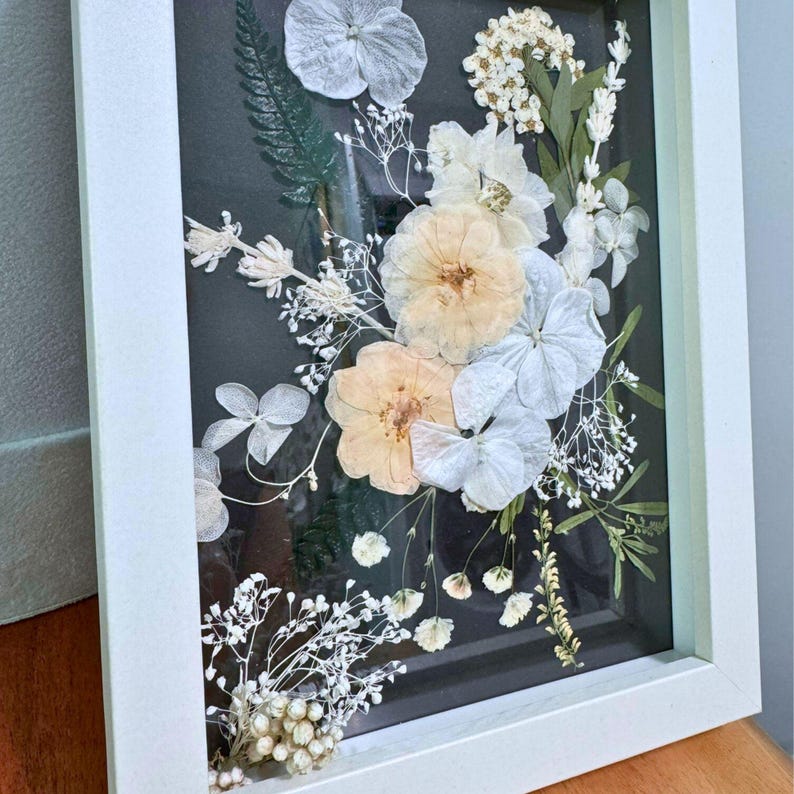 May include: A rectangular white frame containing a pressed floral arrangement on a dark background. The display features dried white and pale peach flowers, including hydrangeas, roses, baby's breath, and green fern leaves arranged vertically.