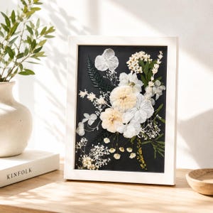 May include: A rectangular white frame containing a pressed flower arrangement on a black background. The composition features white and cream-colored dried blossoms, delicate baby's breath, and dark green fern leaves arranged in a vertical display.