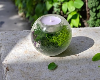 May include: A clear glass orb candle holder with a white candle inside. The orb is filled with green moss and small purple decorative elements. The candle holder sits on a stone surface with green foliage in the background.