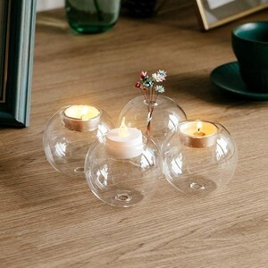 May include: Four clear glass orb candle holders with lit candles and a small vase with flowers. The candle holders are round with a flat base. The candles are small, round, and white. The vase is small and clear.